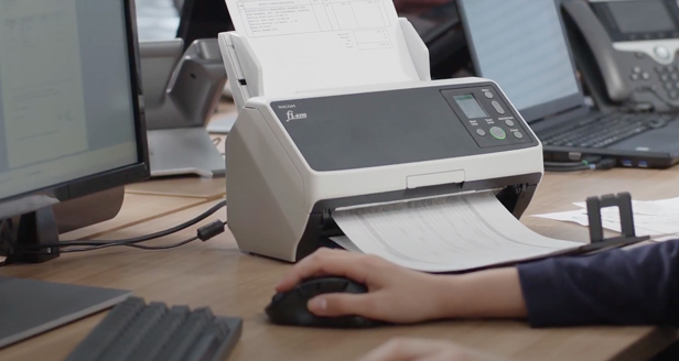 Nahaufnahme einer Hand, die eine Computermaus bedient, während ein Ricoh-Fi-Scanner auf dem Schreibtisch steht